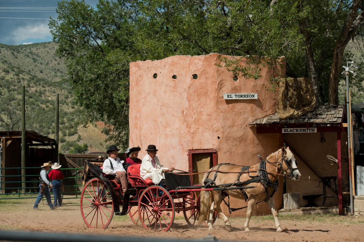 Several people in historical costumes gather near a red horse-drawn carriage in front of "El Torreon," a rustic building, with green hills in the background and other individuals involved in various activities, evoking an old western setting.