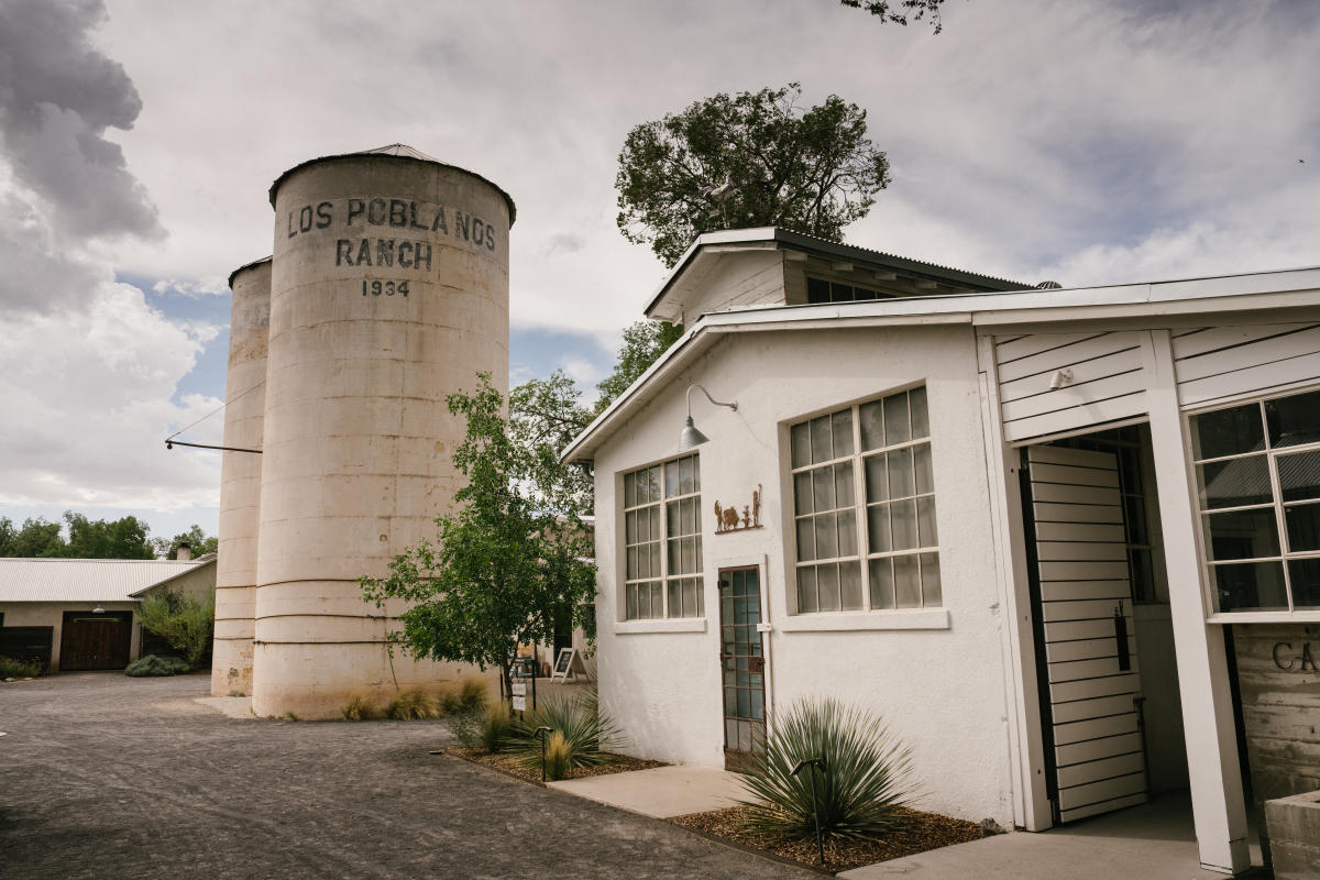 A rustic scene at Los Poblanos Ranch featuring a large silo with the name and year "Los Poblanos Ranch 1934" written on it. Adjacent to the silo, there is a white, two-story building with a tree visible behind it. The area has a desert-like appearance with sparse vegetation.