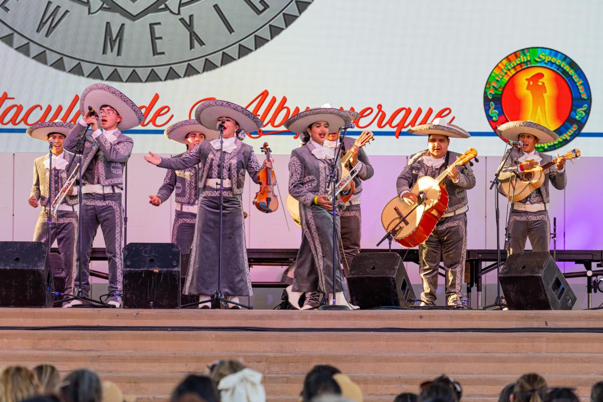 A mariachi band performing on stage, dressed in traditional charro outfits with wide-brimmed sombreros. The group includes violinists, guitarists, and a trumpeter.