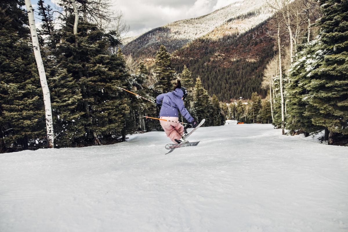 A skier wearing colorful jacket and pants jumps on skis down a snowy slope surrounded by pine trees and mountains.