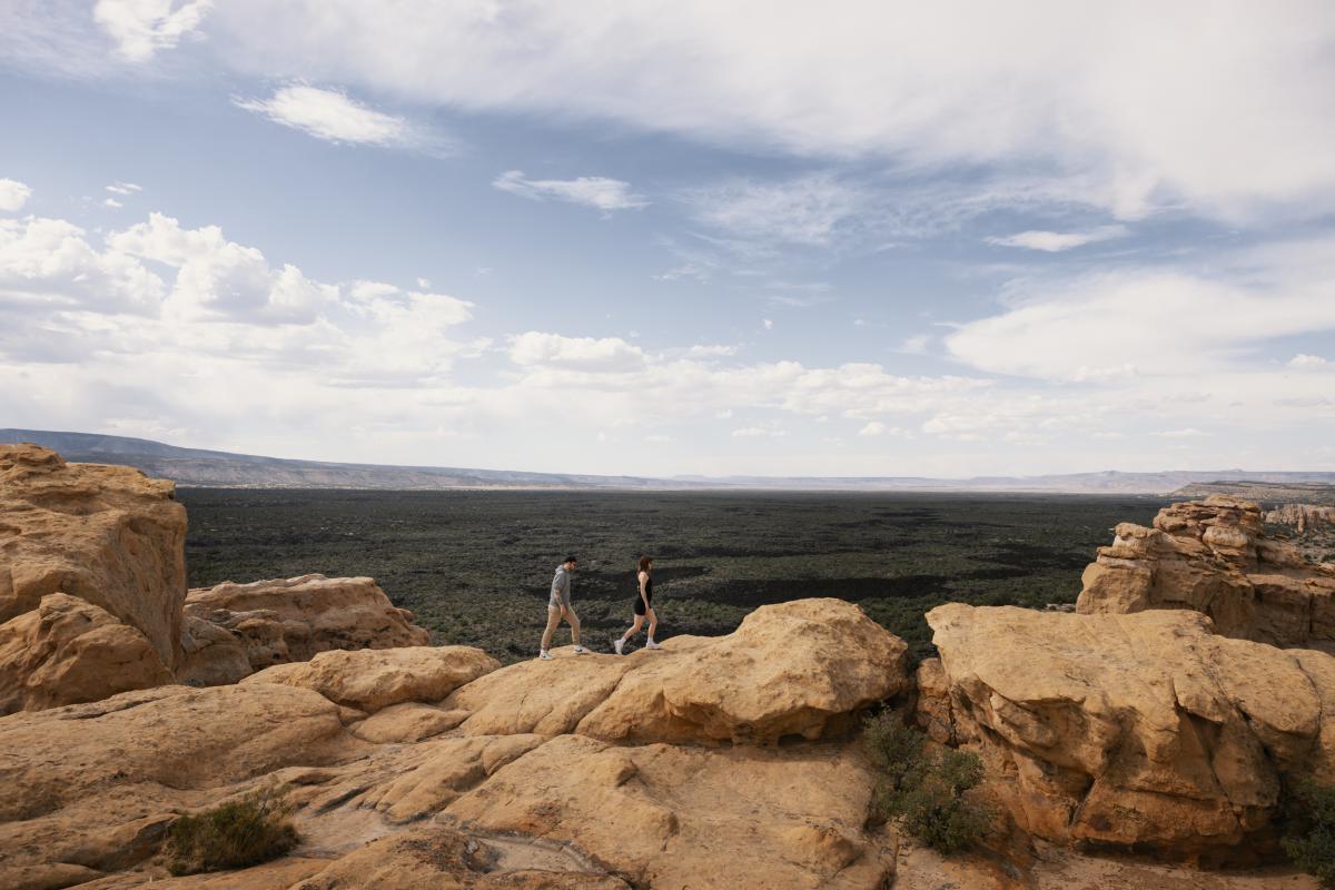 Two people hiking on rocky terrain in a vast desert landscape under a cloudy sky.
