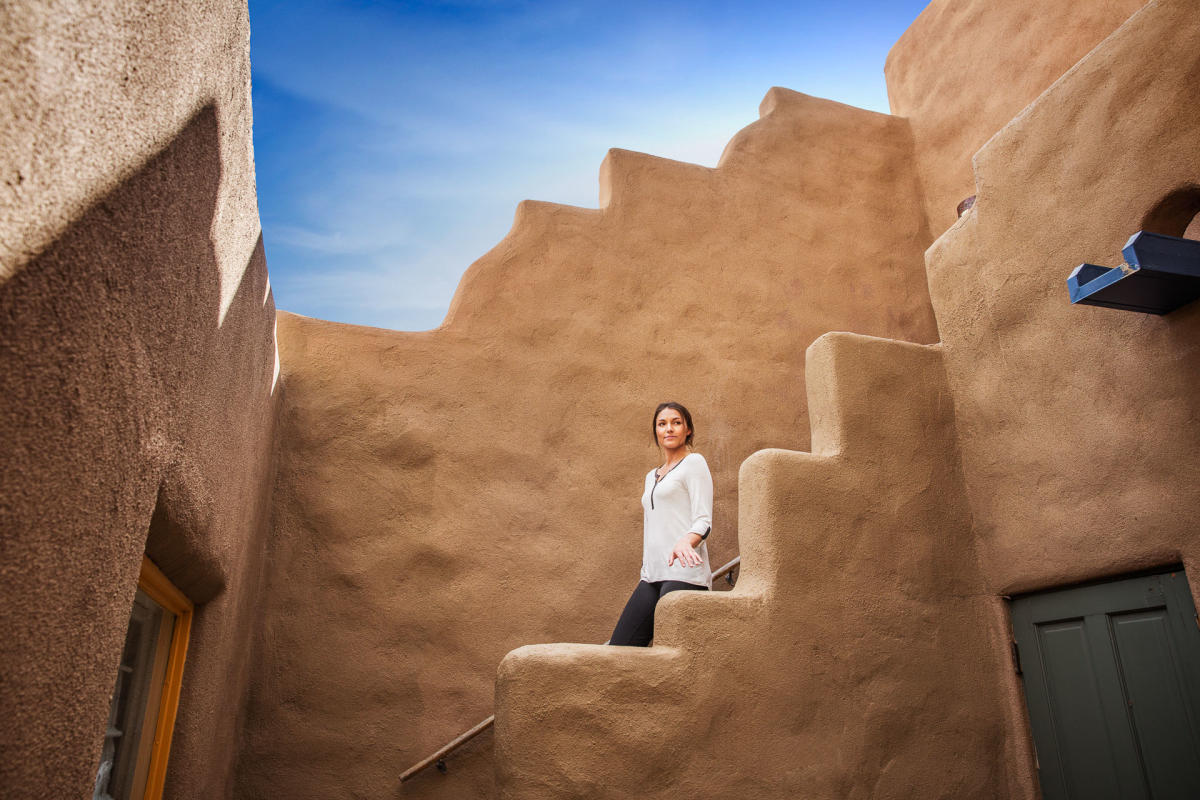 A person stands on a staircase in an adobe-style building under a clear blue sky. The building features smooth, curving walls, typical of Southwestern architecture.