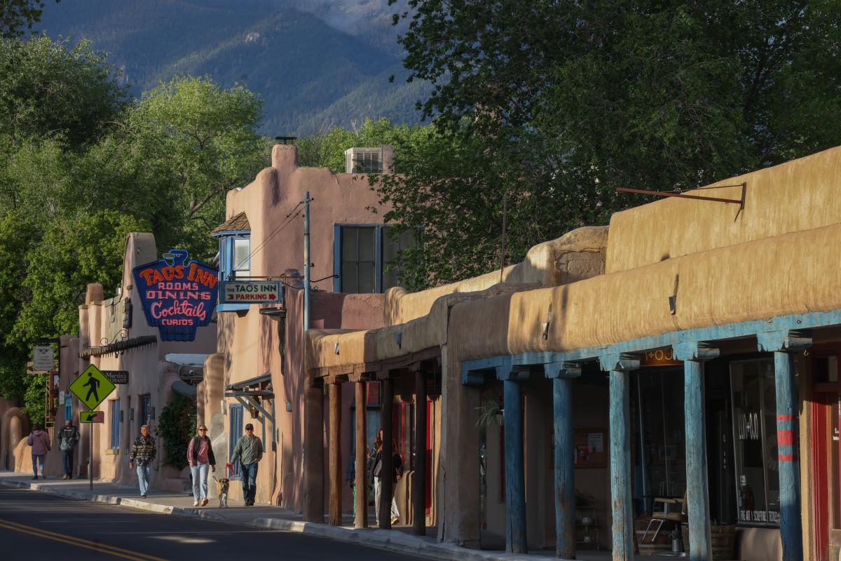 View of a street in Taos, New Mexico, lined with adobe-style buildings. Signs for local businesses, including the 'Taos Inn', are visible.