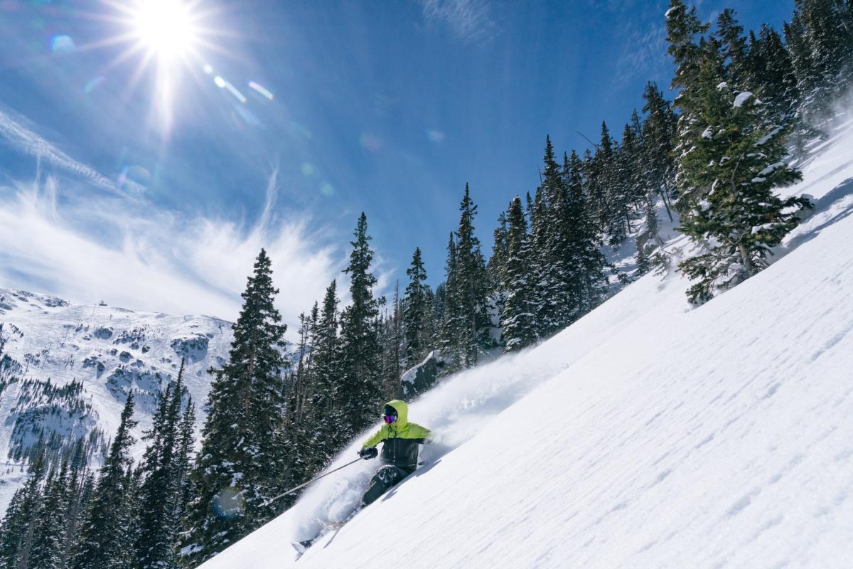 A person skiing down a snowy slope with a backdrop of densely forested mountains under a clear blue sky.