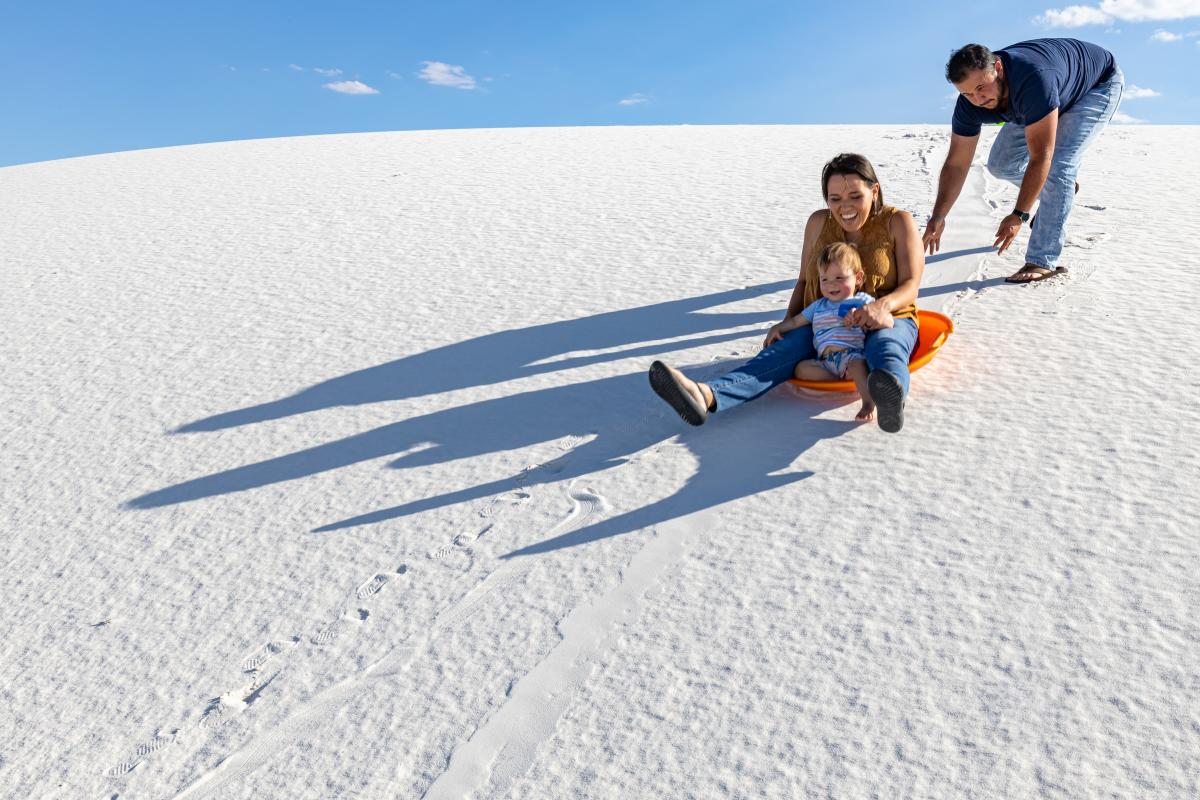 A woman and her child slide down a white sand embankment on an orange sled with a push from another person