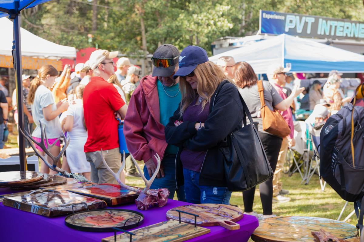 Two people examining artwork at a booth at an outdoor community festival, with other attendees and booths visible in the background.