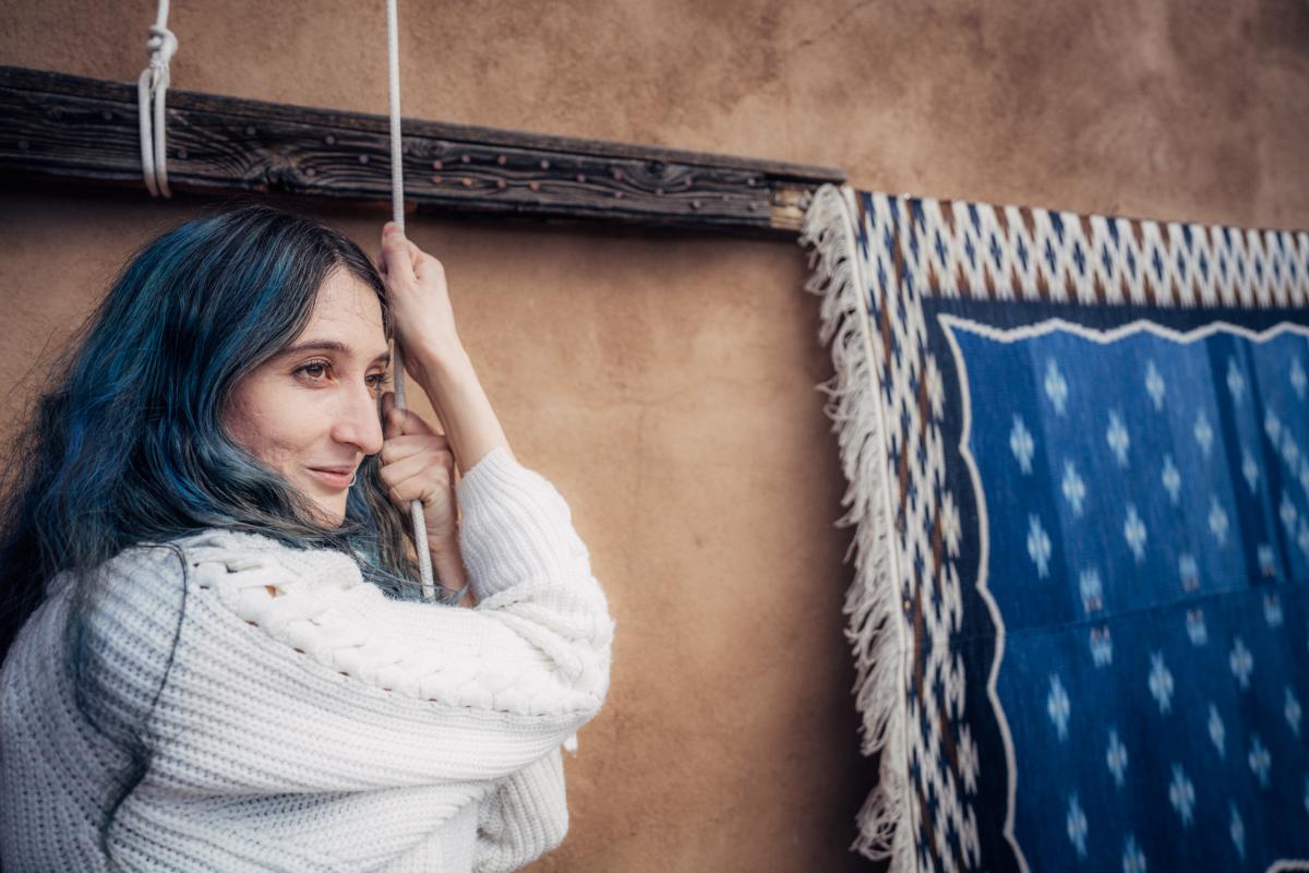 Person with blue hair wearing a white sweater, leaning on a wooden beam beside a hanging blue textile with a geometric pattern.