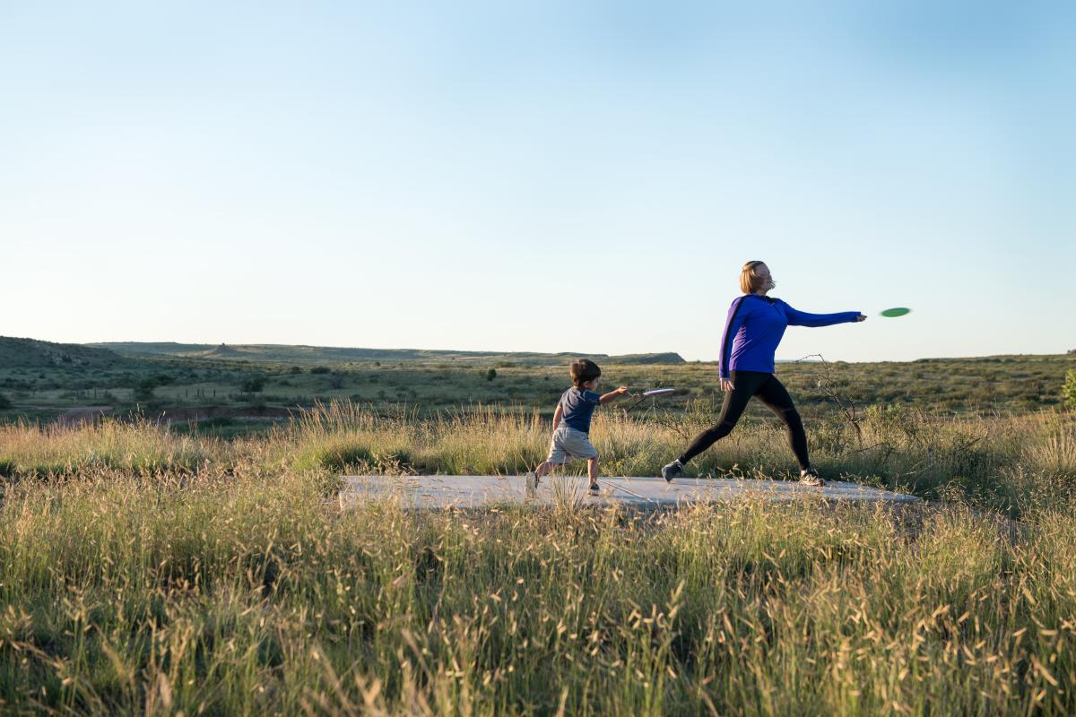 Two people playing disc golf in a grassy field with a scenic backdrop.