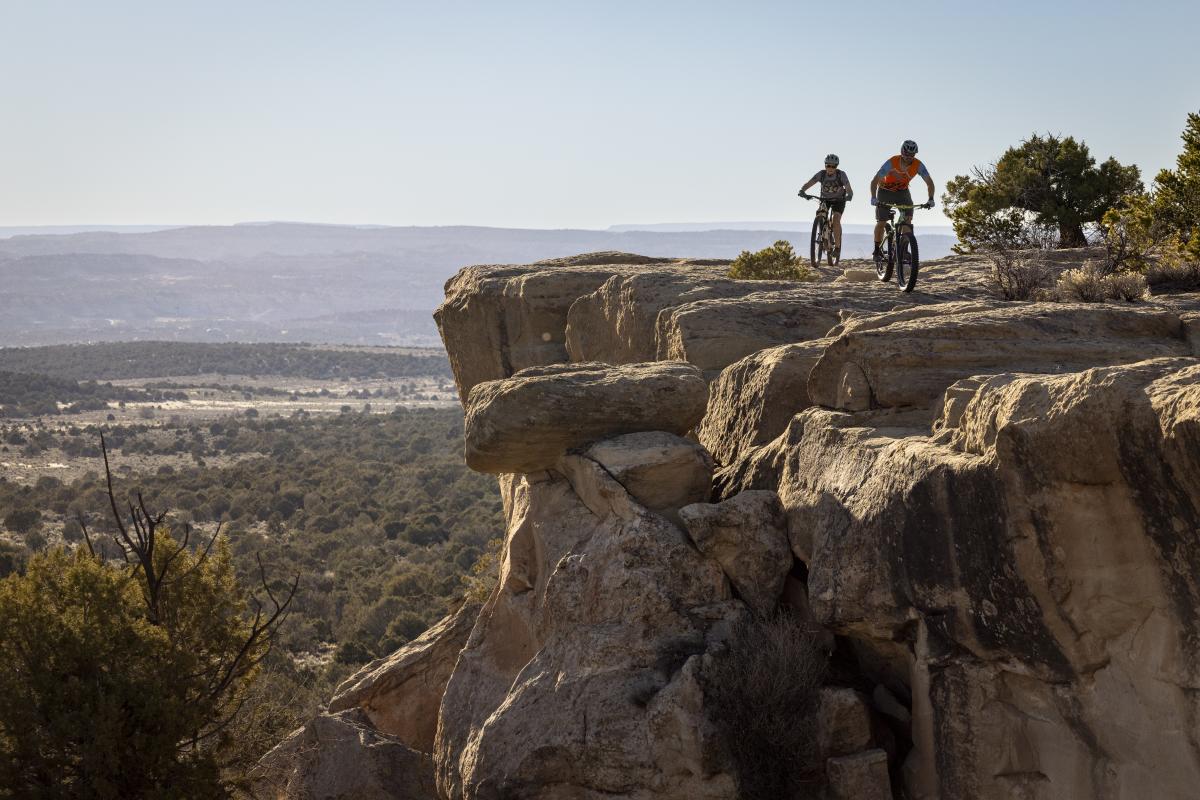 Two cyclists riding mountain bikes on a rocky overlook, enjoying a scenic view of a vast valley.