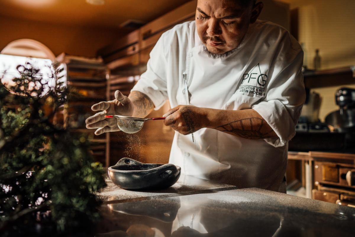 Chef Justin Pinoche delicately spreads powdered spices from a sieve onto a small clay dish standing in a dim but warmly lit kitchen