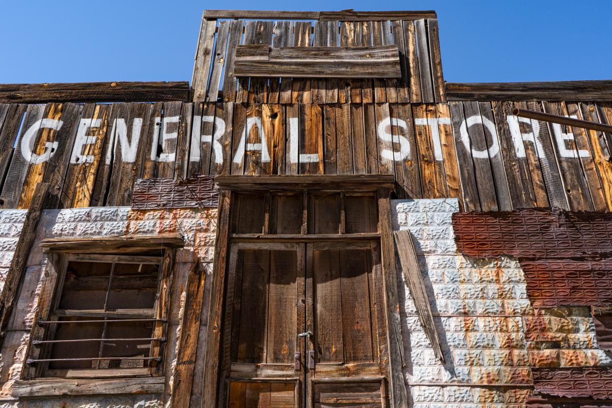 Front view of an old, weathered wooden building labeled 'General Store' with a boarded-up window and a faded, peeling paint facade under a clear blue sky.
