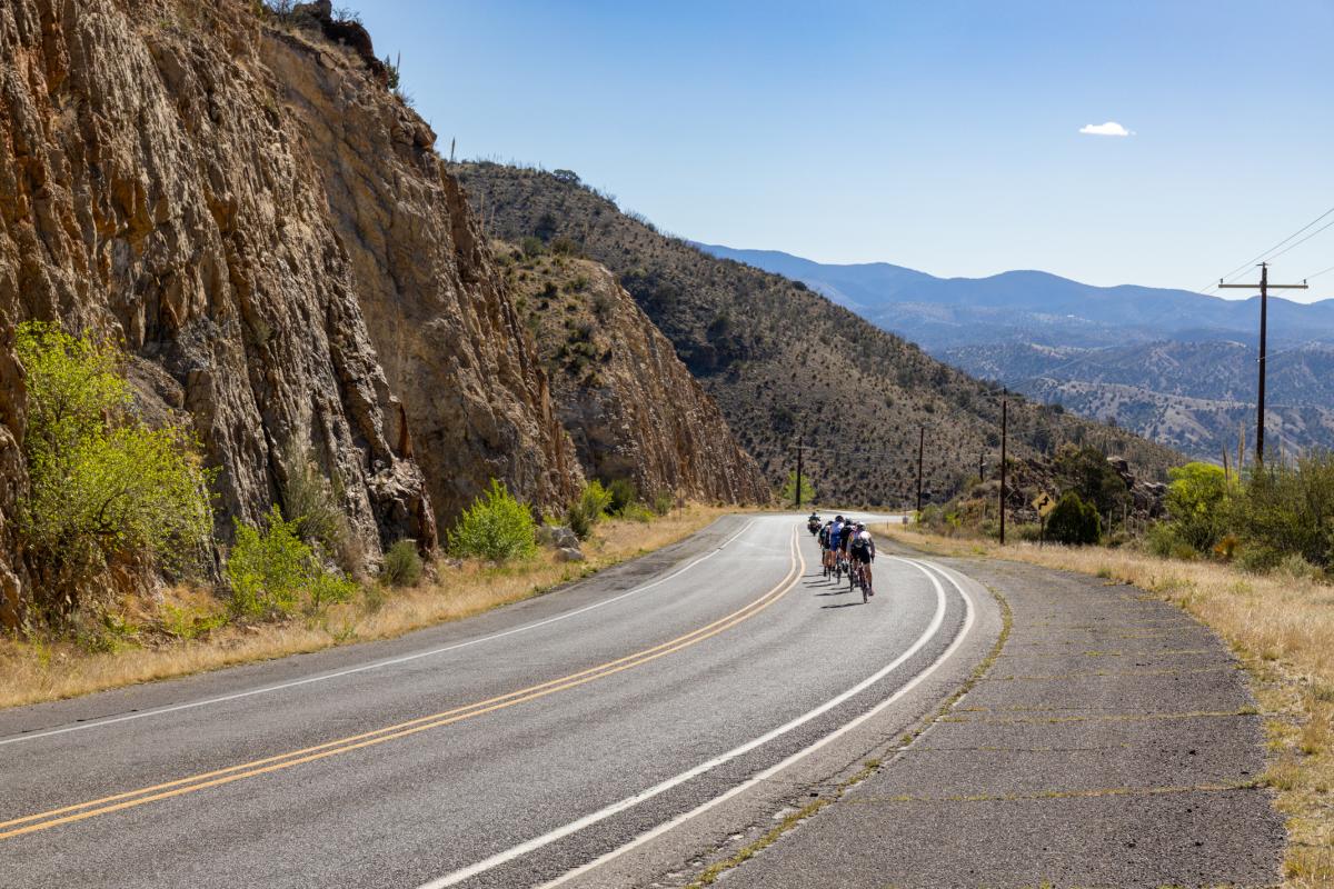 Image showing a group of cyclists riding along a winding road flanked by rocky cliffs on one side and a panoramic view of distant hills under a clear sky.