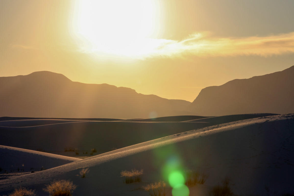 Sunset over the sand dunes at White Sands National Park, with the sun casting a warm glow and creating long shadows on the textured sands.
