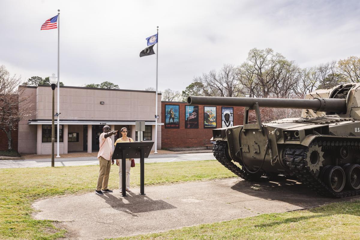 A man points at an informational sign near a tank outside the Virginia War Museum, with military murals on the building in Newport News