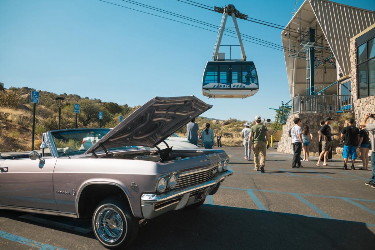 Convertible with hood open in parking lot, gondola labeled “Sandia Peak” above, people walking toward cable car station.