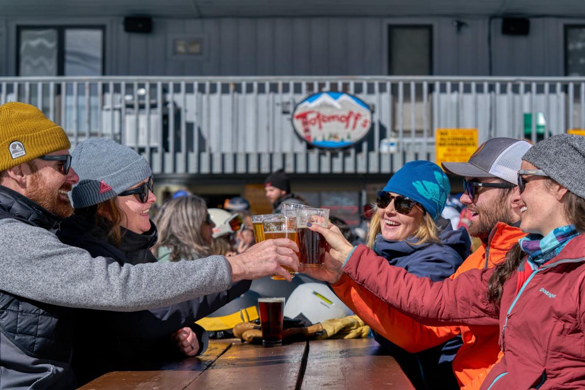 A group of six people wearing winter jackets and beanies cheerfully clink beer glasses at an outdoor table on a sunny day. A ski lodge sign is visible in the background.