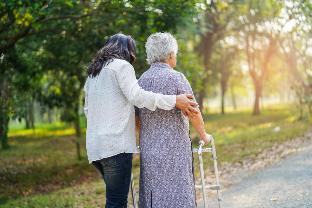A young woman gently assists an elderly woman with a walker, walking through a sunlit park.