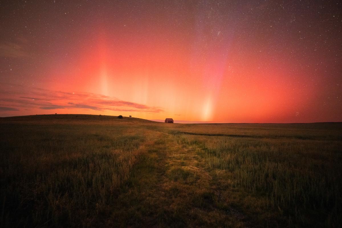 A barn sits on the horizon of a grassy field beneath a vibrant red aurora borealis.