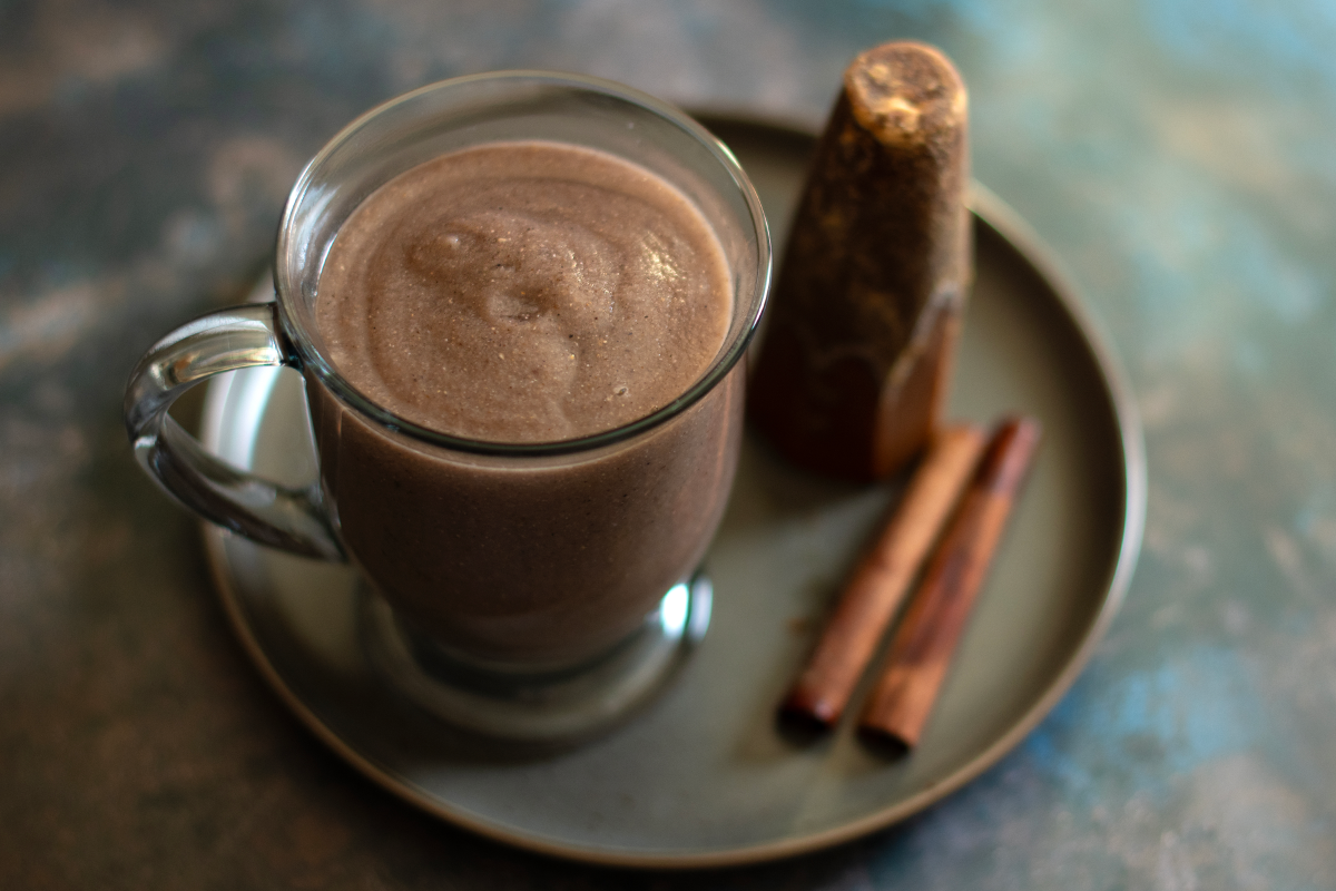 A glass mug of a thick blue corn atole sits on a plate with a cone of piloncillo and two cinnamon sticks.