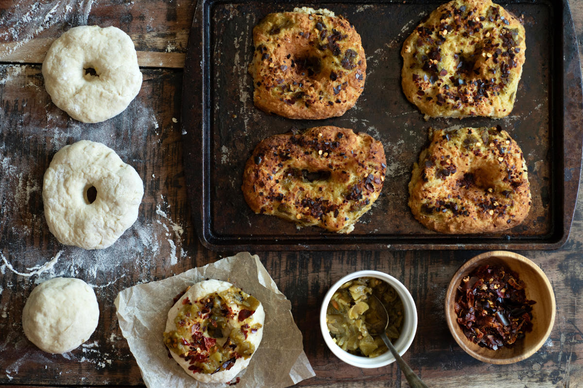 An overhead shot of a wooden table with ingredients to make green and red chile bagels.