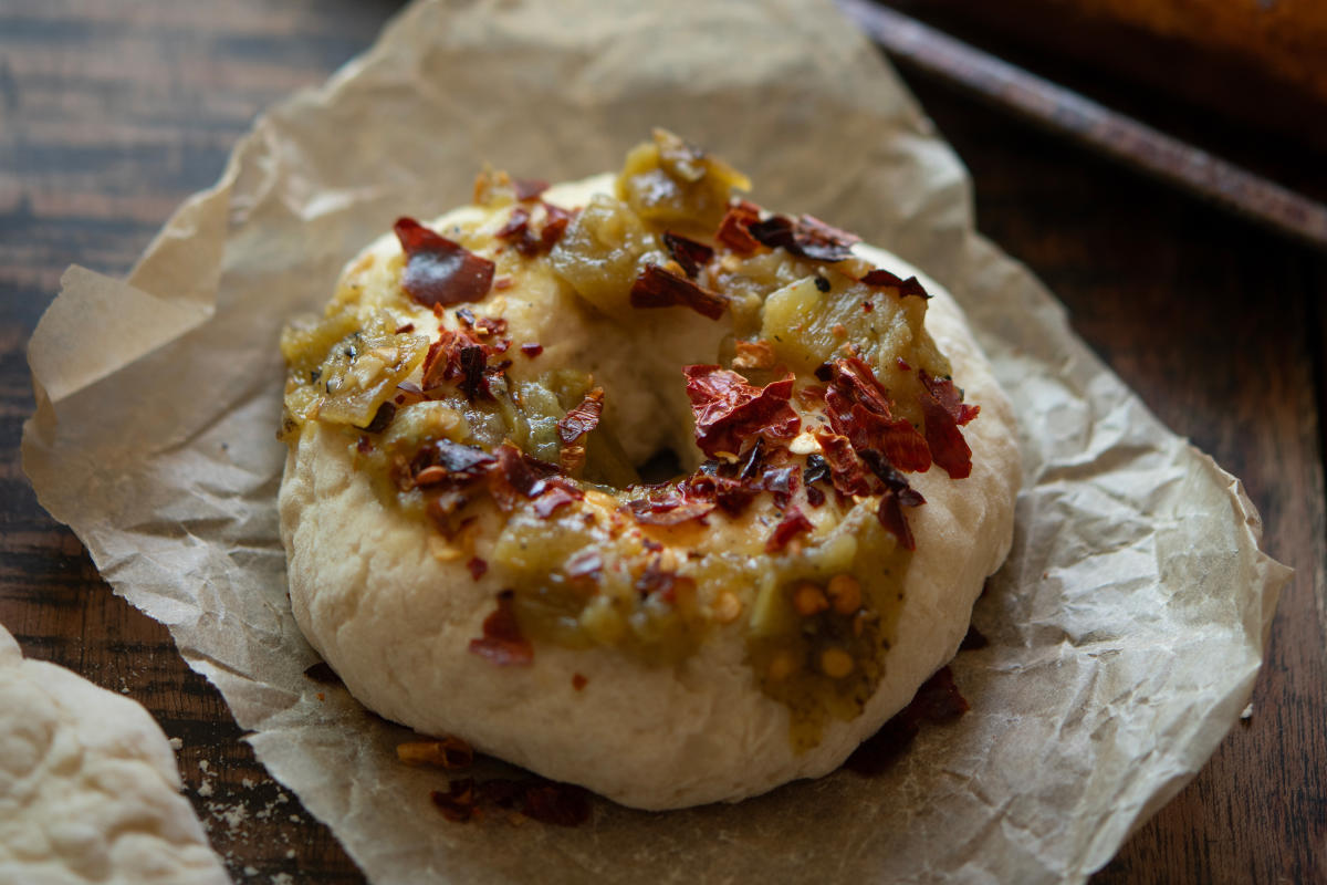 A bagel topped with chopped green chiles and red pepper flakes rests on brown parchment paper.