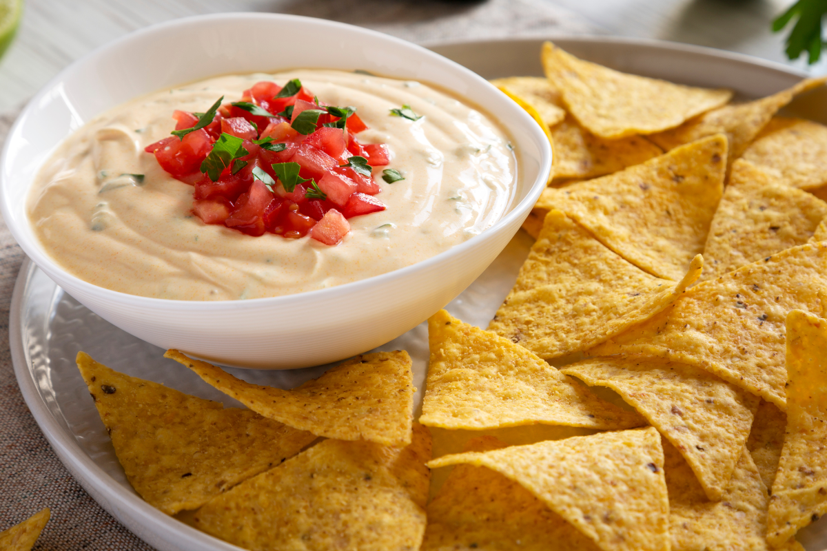 A bowl of chile con queso topped with diced tomatoes and surrounded by tortilla chips.
