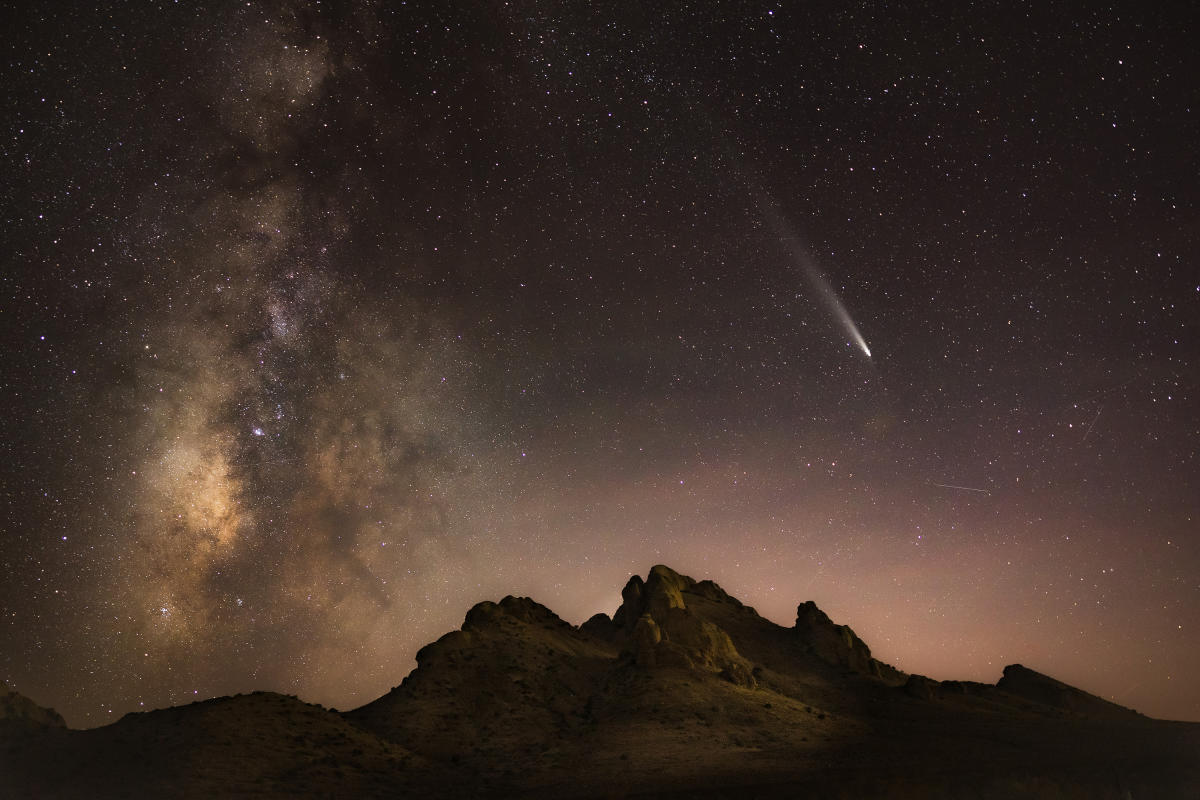 Starry night sky over a silhouetted mountain range, featuring a bright comet with a long tail and the Milky Way galaxy visible.