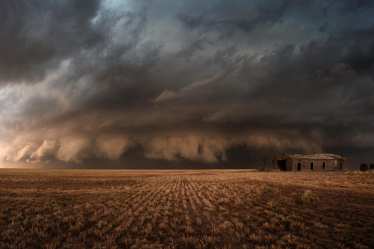 A massive, dark storm cloud hovers over an abandoned farmhouse and a barren field.