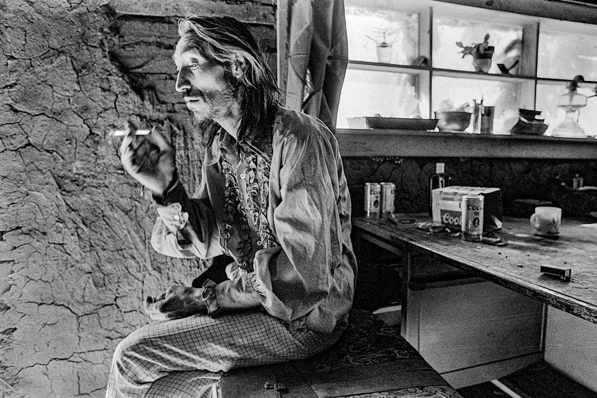 Black and white portrait of a long-haired man sitting indoors beside a rough adobe wall, holding a cigarette.