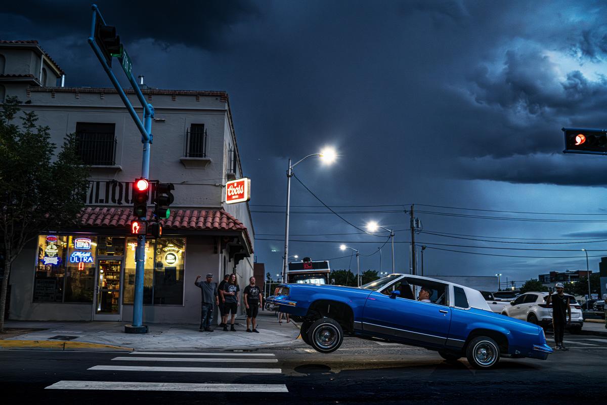 Blue lowrider car lifting its front wheels at a street corner under dark stormy skies.
