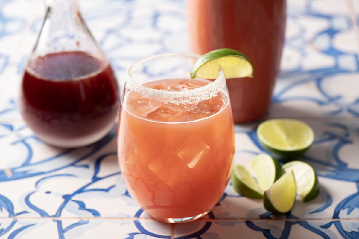 A prickly pear margarita glass with ice and a sugared rim, garnished with a lime slice, sits on a patterned tile table. Nearby are lime wedges and a pitcher of prickly pear syrup.