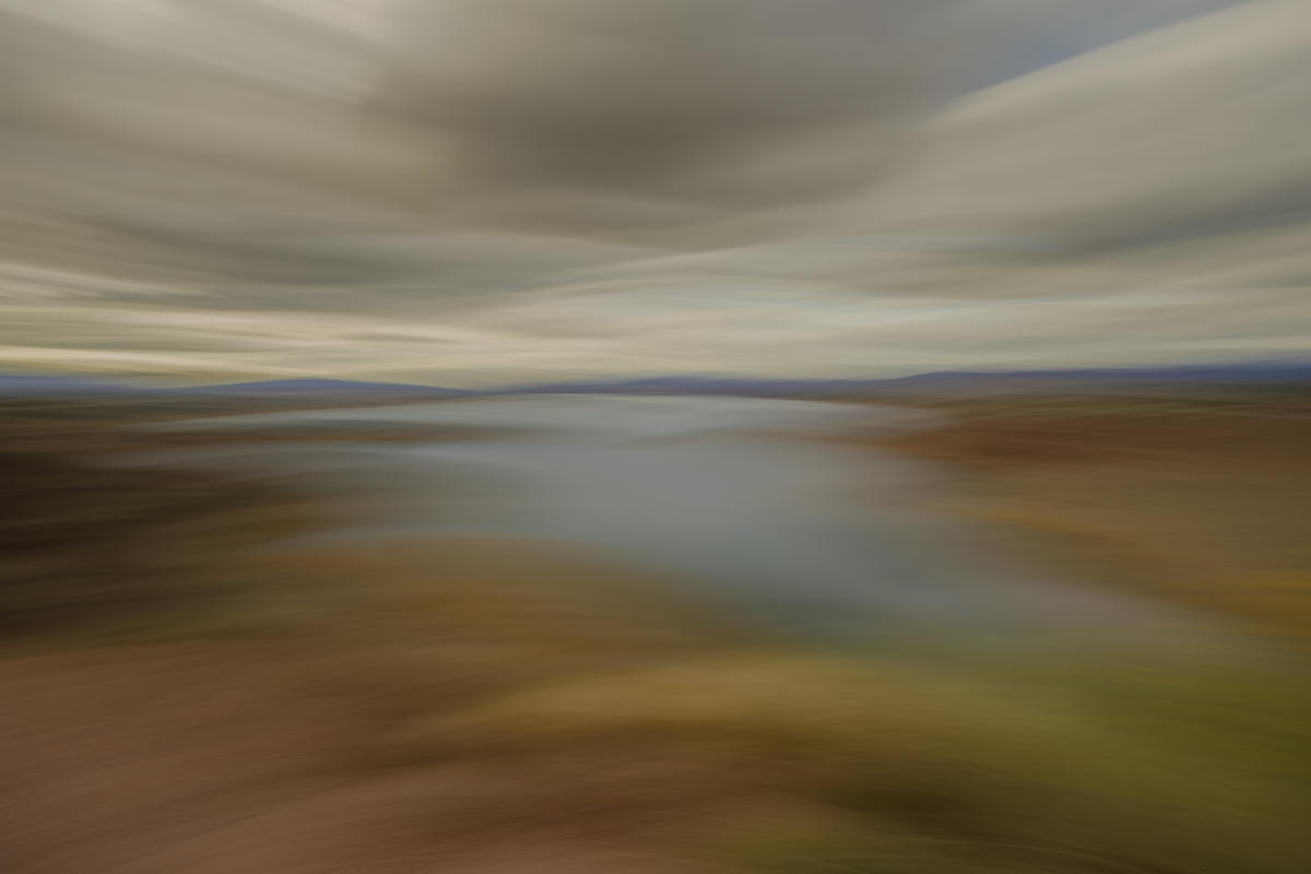 A dynamic landscape with blurred motion effect; a river runs through earthy tones under an expansive, cloudy sky.