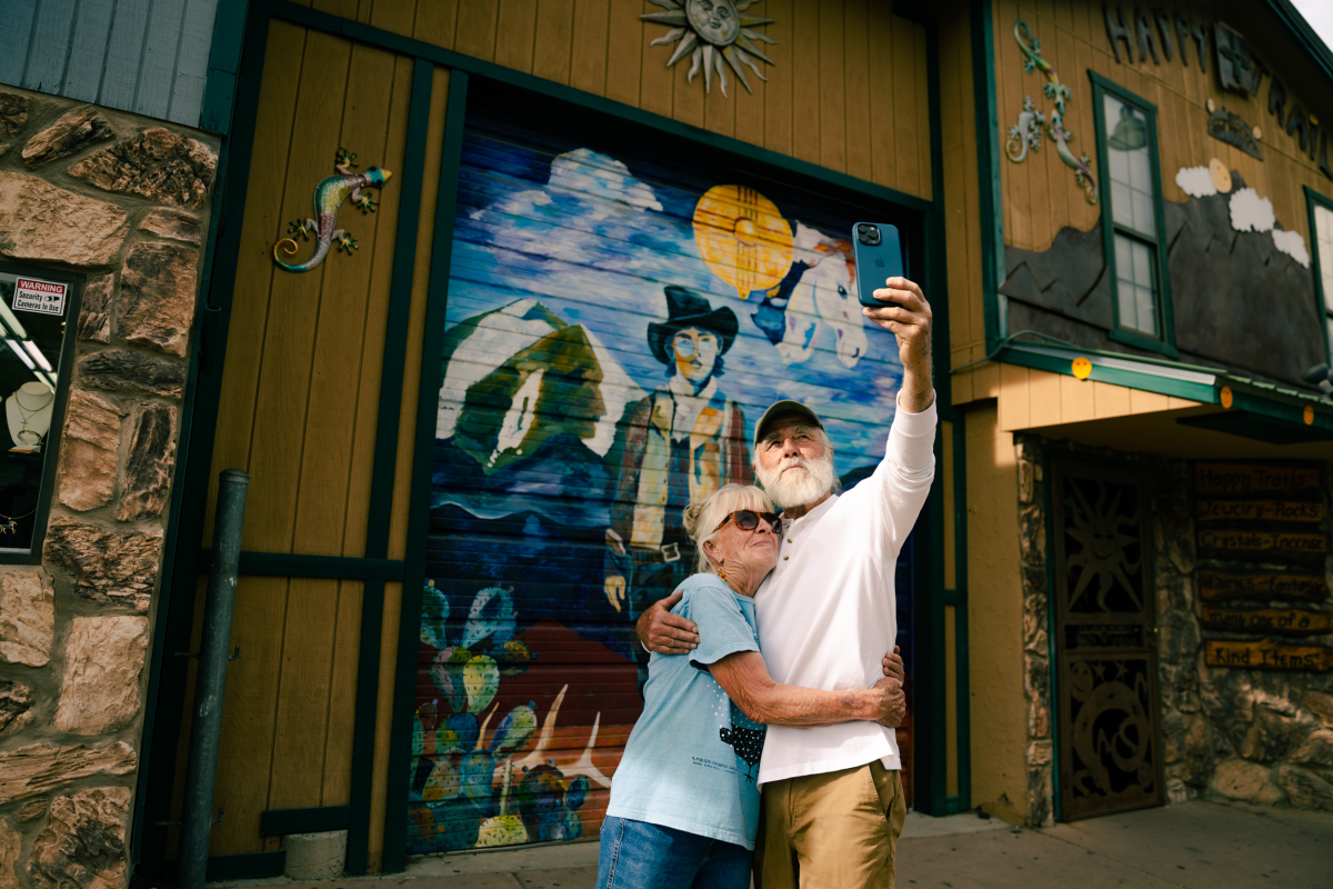 A couple hugs in front of a mural while taking a selfie.
