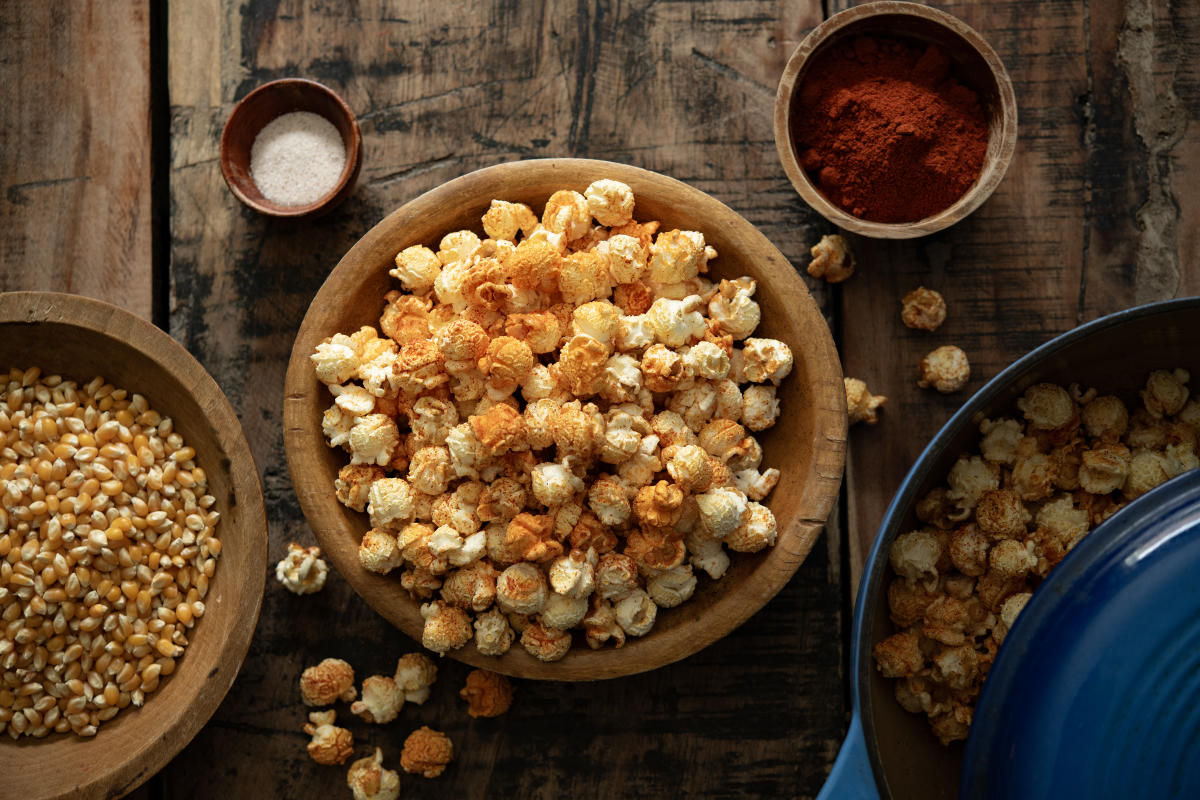 A close-up overhead view shows a wooden table filled with bowls of popped popcorn, unpopped kernels, and seasonings like salt and red chili powder.