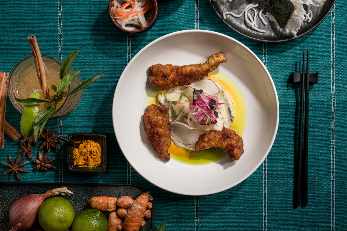 A white plate with crispy fried chicken topped with radish slices and microgreens, set against a teal cloth. Ingredients and chopsticks surround it.