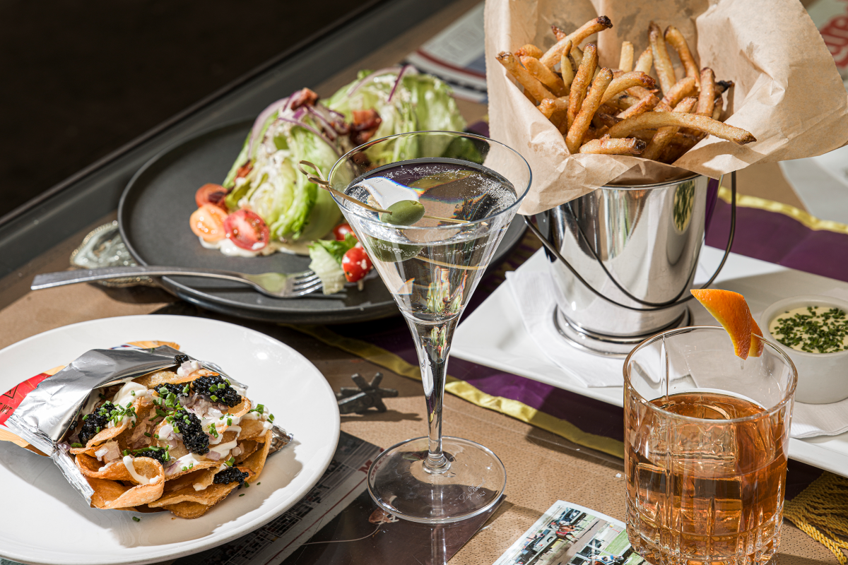 A gourmet spread featuring a martini with an olive, a bucket of crispy fries, a wedge salad with tomatoes, and fancy nachos.