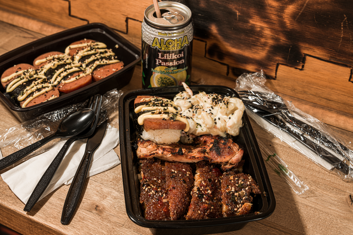 A takeout container with Spam musubi, glazed chicken, macaroni salad, and another with sliced spam topped with sauce. A can of Aloha Lilikoi Passion and utensils are nearby.