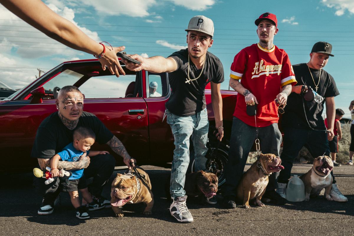 A group of men with their dogs stands in front of a lowrider, with an arm holding a phone entering the corner of the frame, capturing a spontaneous moment at Duke Day.