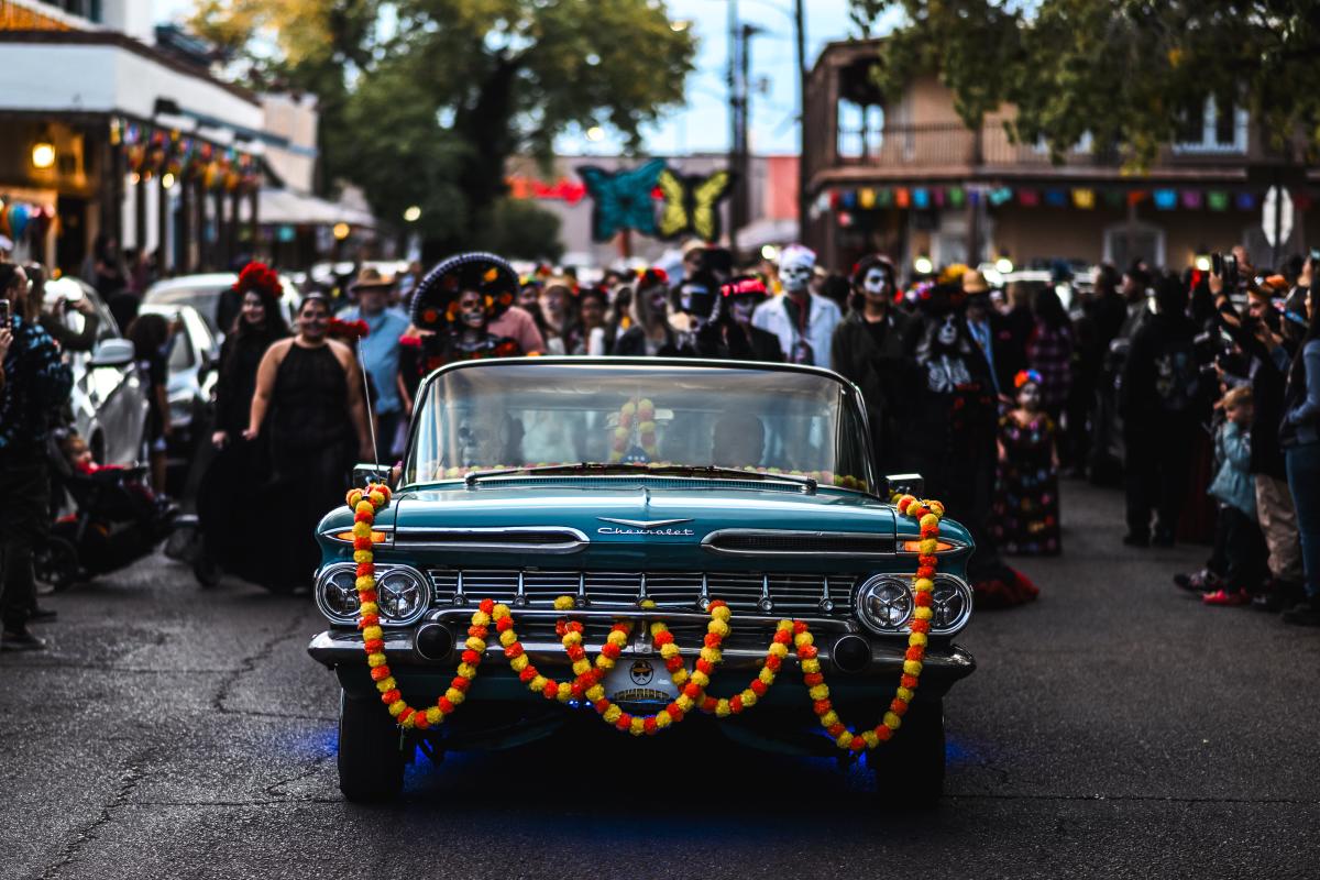 A man drives his 1959 Chevrolet El Camino at the front of the Día de los Muertos Parade in Old Town, surrounded by parade activity.