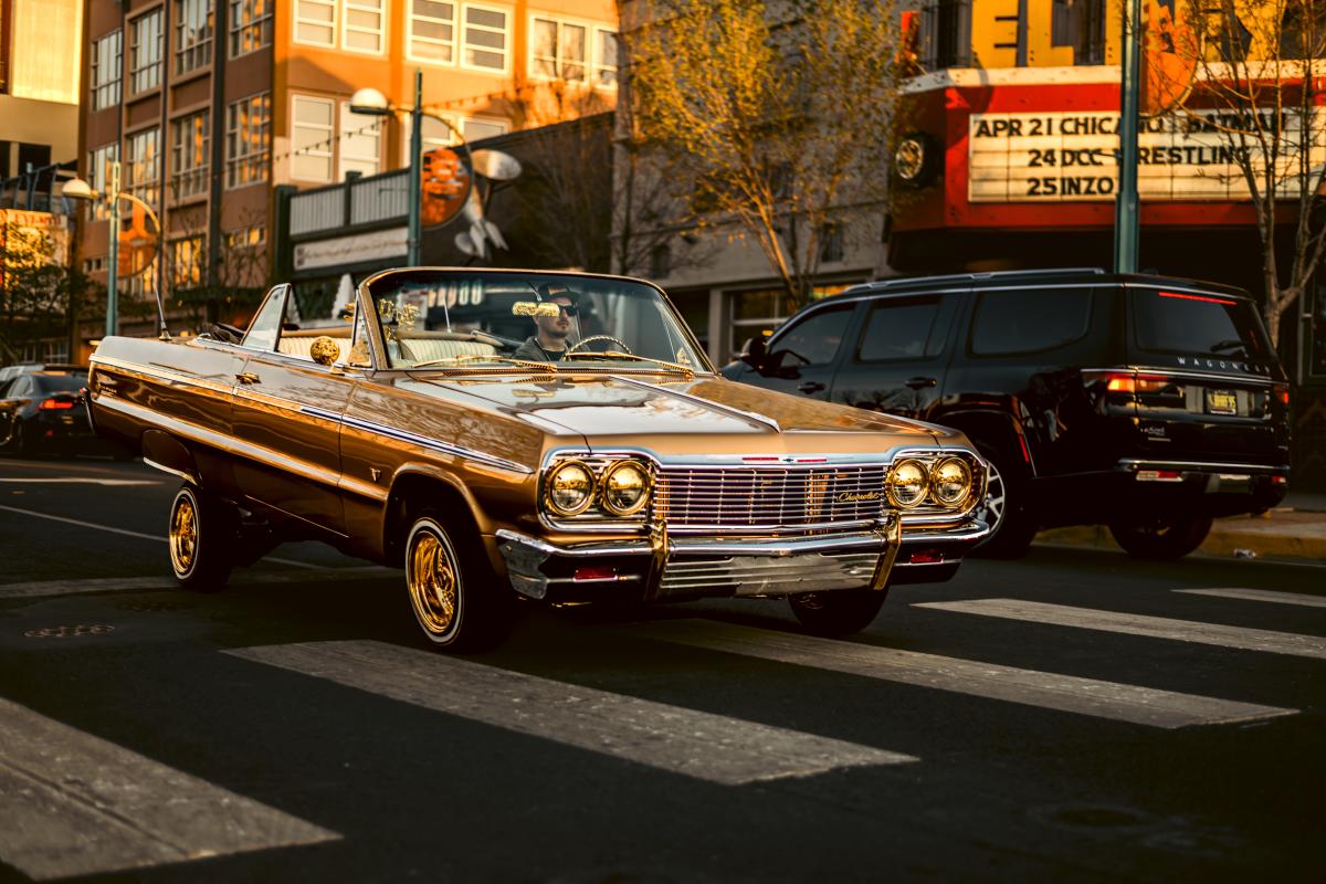 A gold Chevrolet Impala cruises through downtown Albuquerque in warm evening light, the paint glowing as it catches the sun.