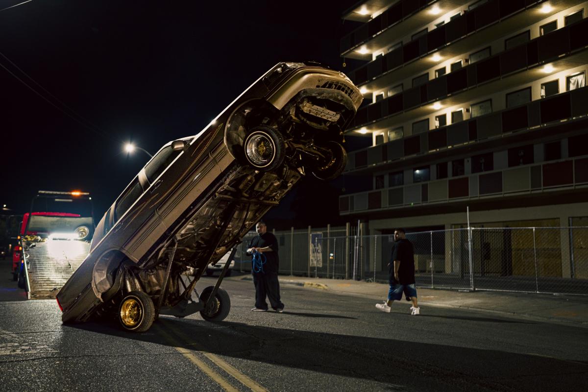 A nighttime scene where a lowrider hopper is being operated, framed by dim streetlights.