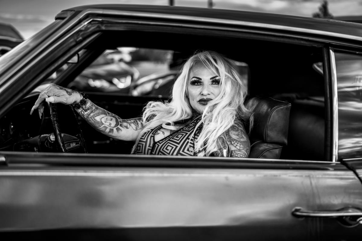 A portrait of Mabel Chavez in a lowrider, dressed in stylish clothing and looking confidently into the camera.