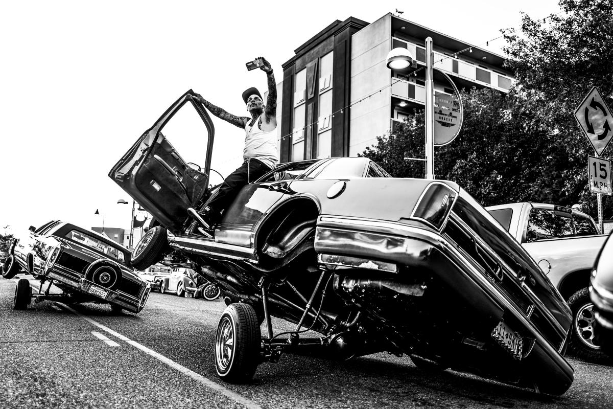 Sonny steps out of his Oldsmobile into a crowded street full of lowriders during Sparky’s Memorial Cruise, with traffic briefly stopped.