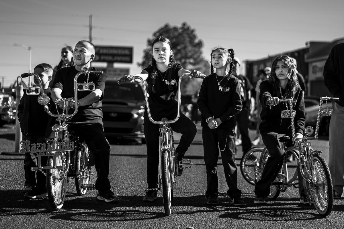 Members of the Razaunida Bike Club display custom lowrider bicycles during a community event, surrounded by people and lowrider cars.