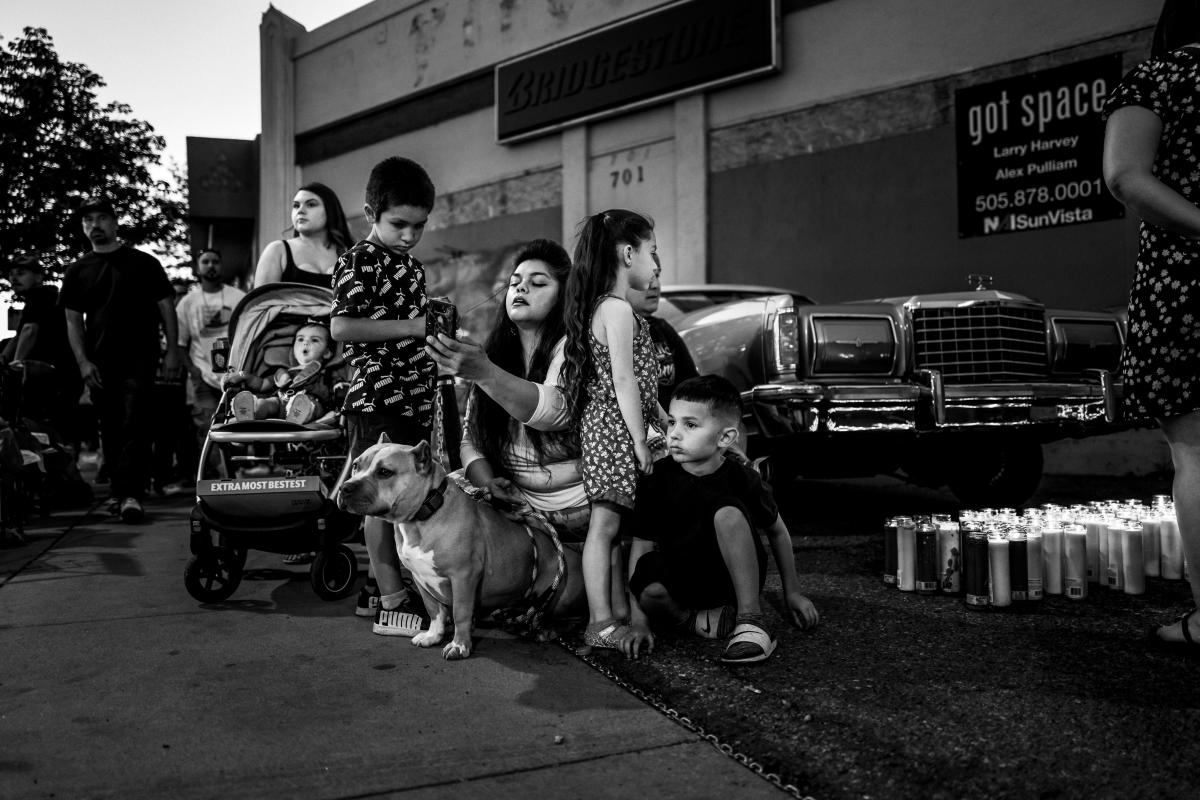 A family sits in front of a memorial display featuring candles and a lowrider belonging to Sparky during a gathering at Seventh and Central.