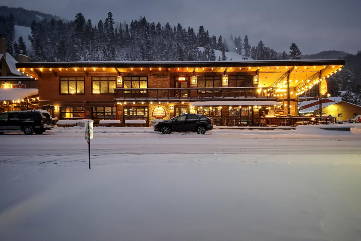 A warmly lit lodge with wood detailing stands amid fresh snowfall. Vehicles are parked in front, and snow-covered trees are visible in the background.