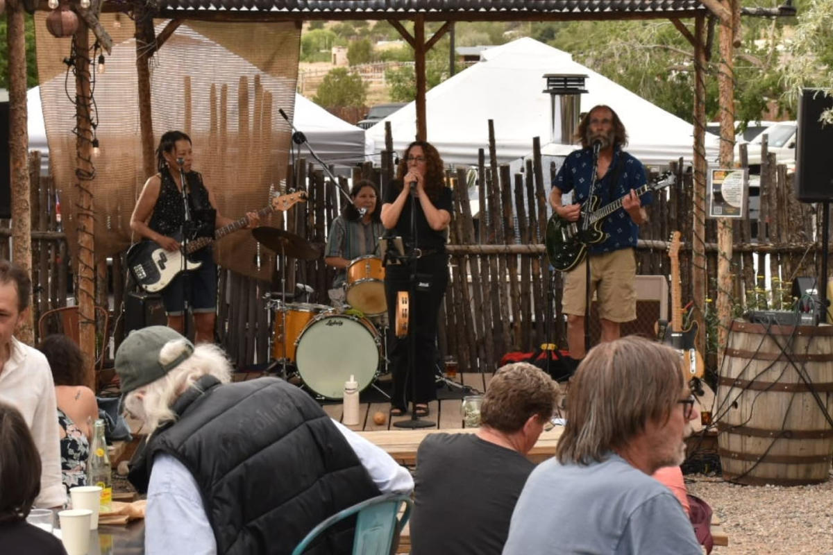 Outdoor band playing on a rustic stage with string lights. Audience seated at tables, enjoying music. Trees and tents visible in the background.