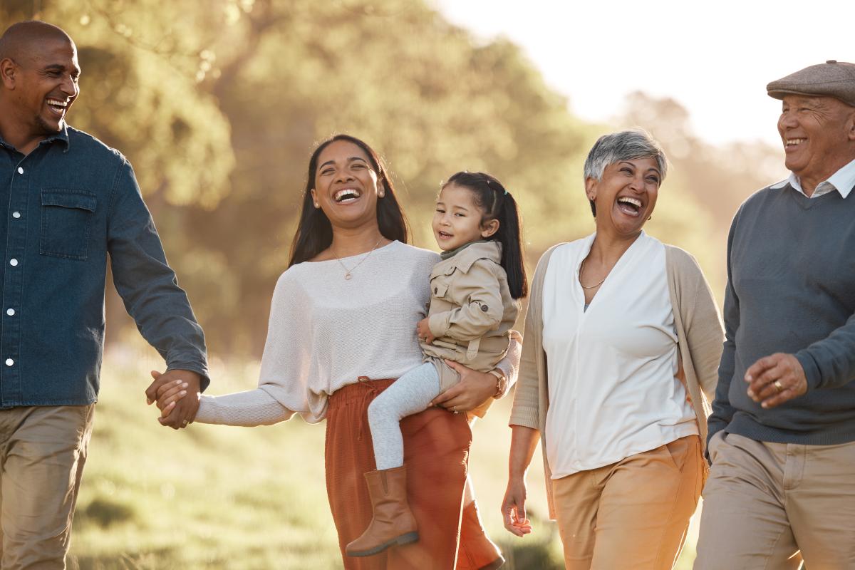 A happy family strolls in a sunlit park, laughing. A woman carries a child, surrounded by an older couple and a man holding her hand.