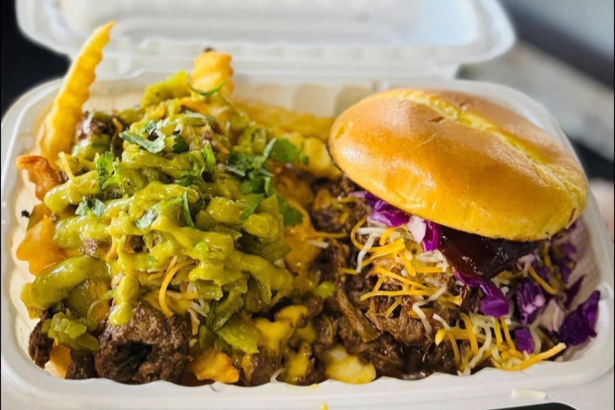 A takeout box with crinkle-cut fries covered in melted cheese, green chili, and herbs sits next to a juicy burger.