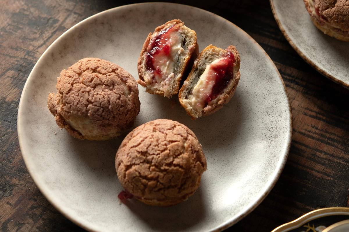 Cream puffs on rustic plates; one puff is halved, revealing creamy filling with red fruit jam.